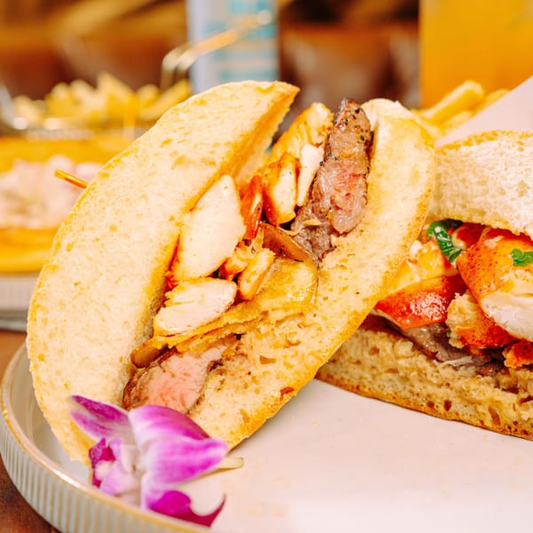 Two gourmet sandwiches, one with steak and chicken, the other with lobster and steak, on a white plate.