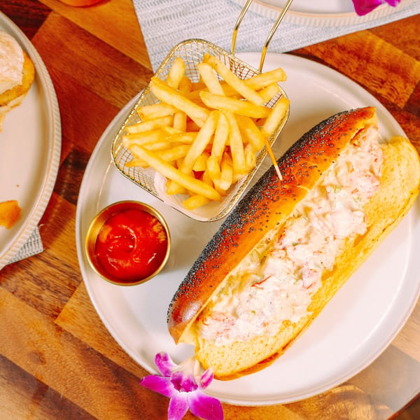 Lobster roll with fries and ketchup on a white plate, garnished with a purple orchid.