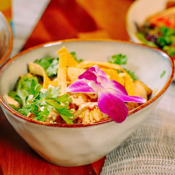 A colorful bowl of rice, cilantro, tortilla strips, and a purple orchid on a wooden table.