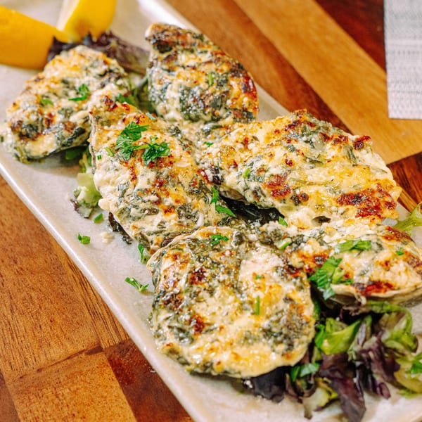 Baked oysters topped with cheese, spinach, and herbs, garnished with lemon wedges on a white platter.
