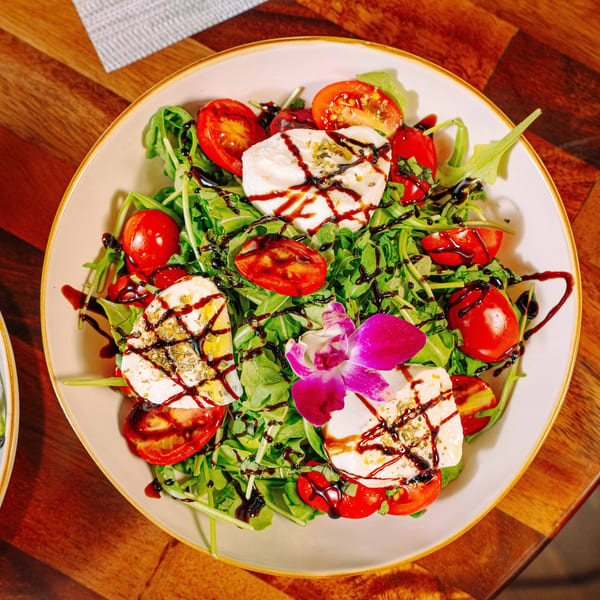 Caprese salad with fresh mozzarella, cherry tomatoes, arugula, balsamic glaze, and an orchid garnish.